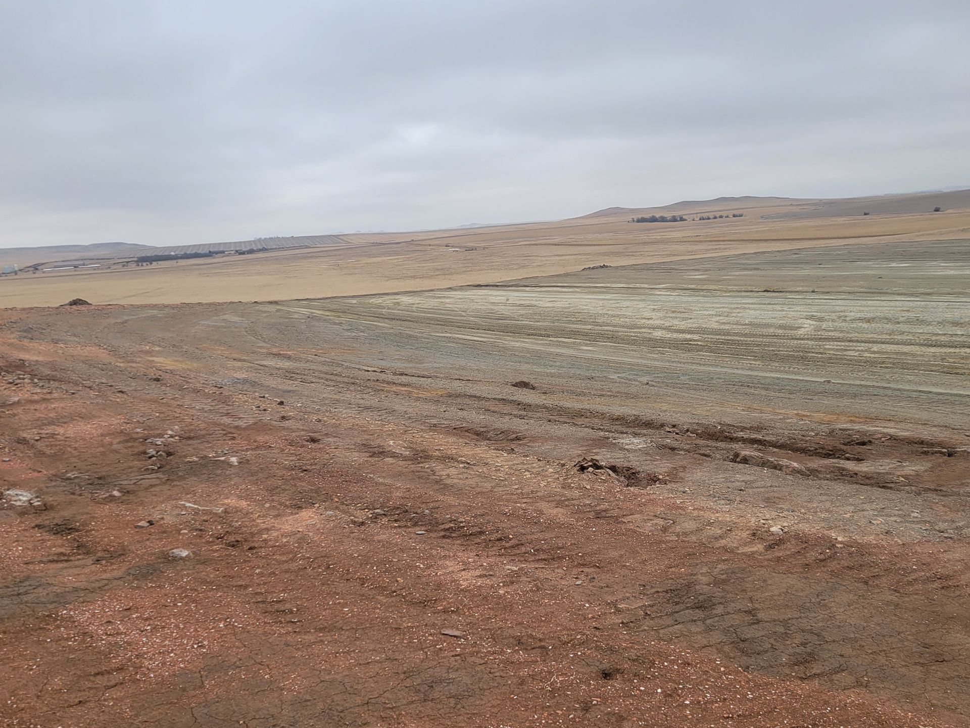 Barren landscape with reddish-brown dirt in the foreground and a tan plain stretching towards the horizon under a cloudy sky.