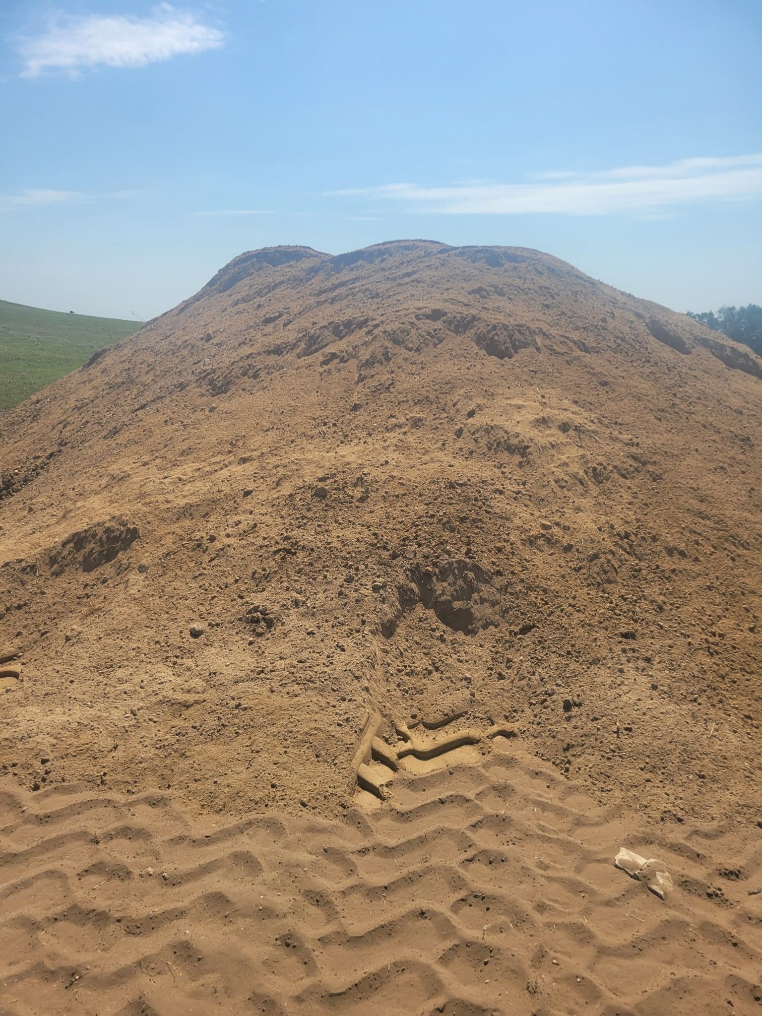 A large mound of tan sand under a bright blue sky, with visible tire tracks in the foreground.