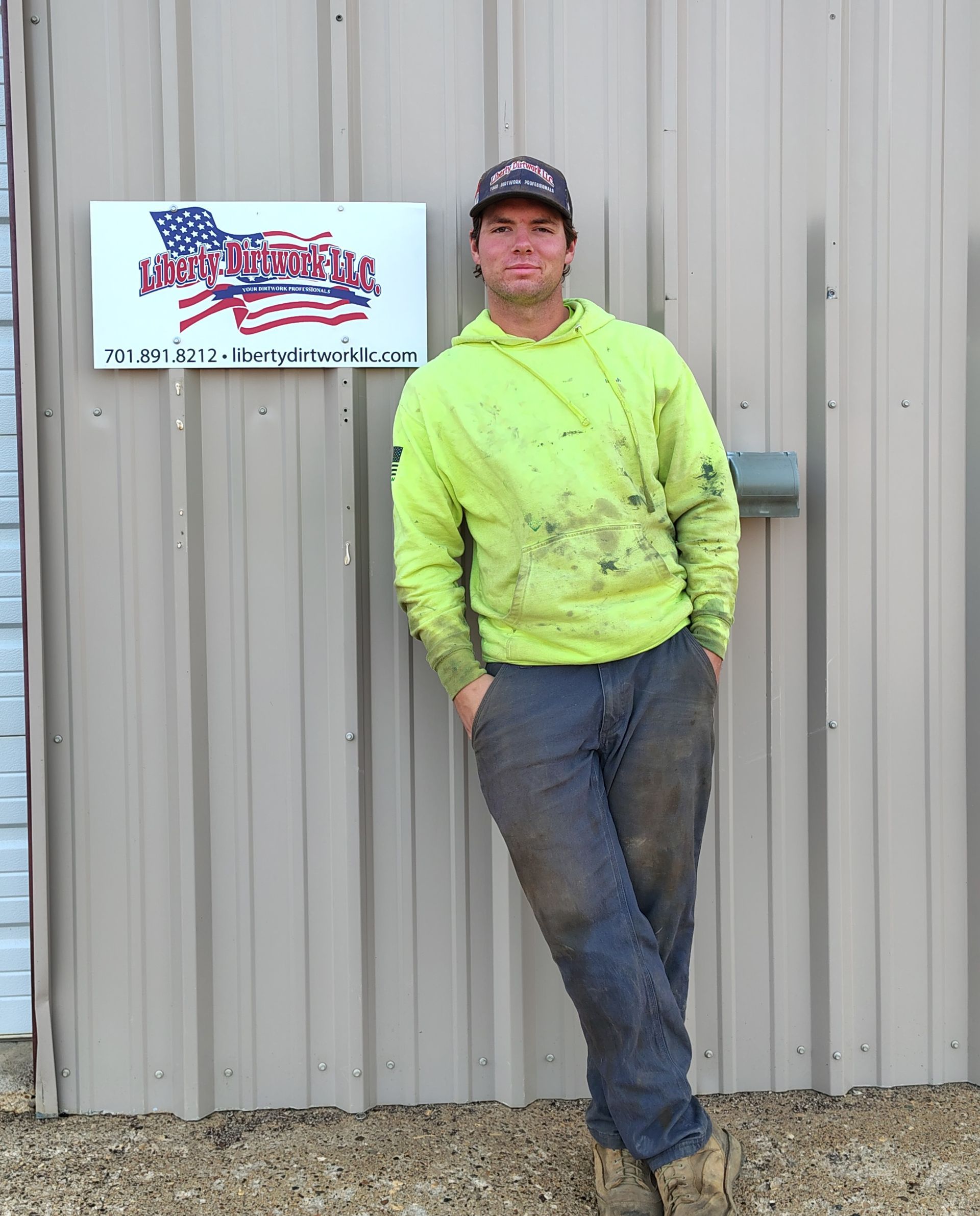 Man in work clothes leaning on building, beside sign reading 