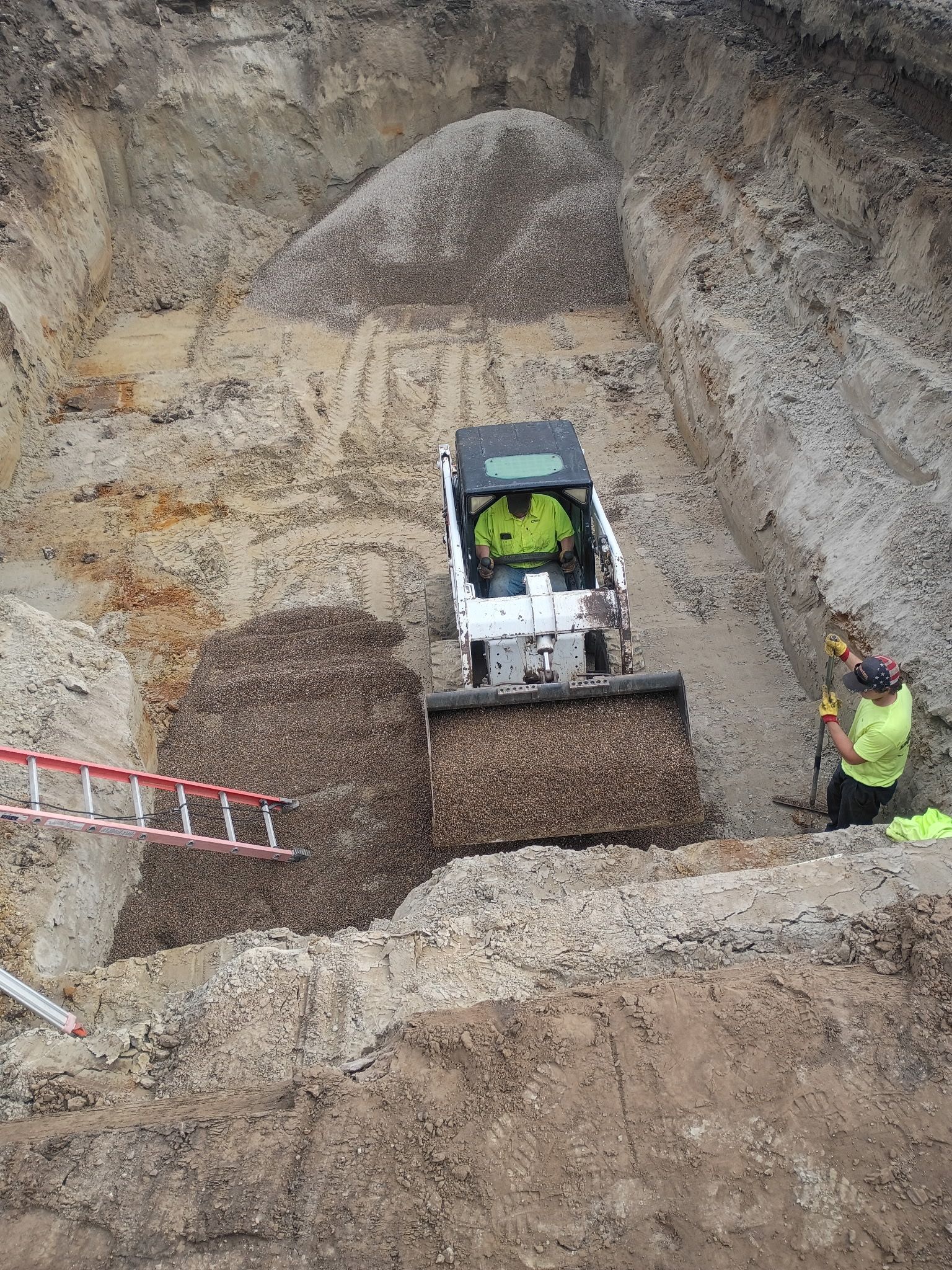 Bobcat loading material into a trench, with a worker and ladder nearby; construction site.