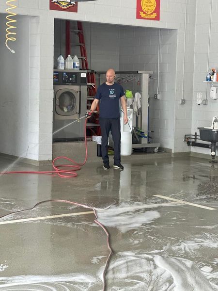 A man sprays a foamy floor in a garage-like space with a red hose; visible washing machine and cleaning supplies.