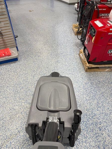 Floor cleaning machine moving down a long aisle in a store with speckled floors and equipment on the right.