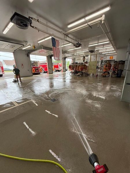 Fire station bay being cleaned with a hose. An American flag hangs inside; fire trucks are parked.