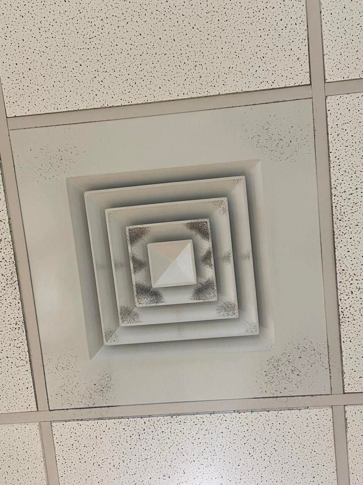 White ceiling vent in a white grid ceiling, with square design and some dark speckling around edges.