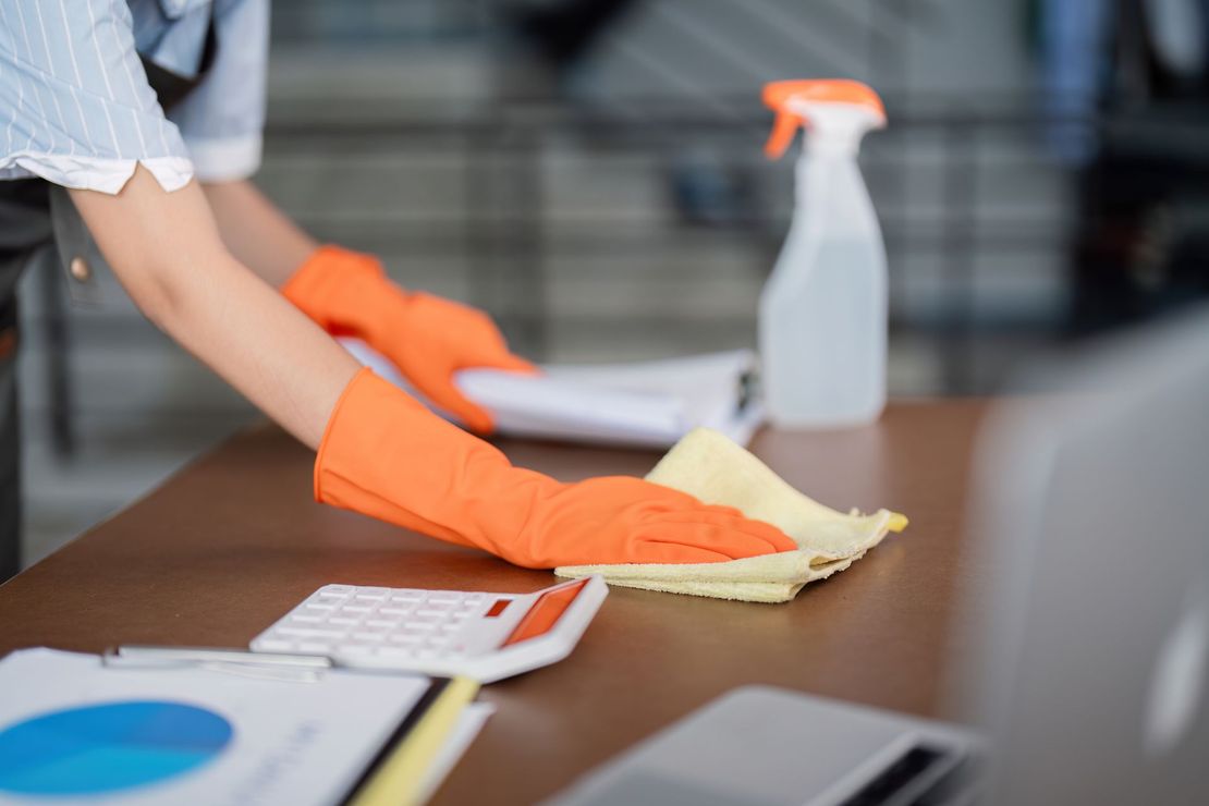Person wearing orange gloves cleaning a wooden desk with a yellow cloth and spray bottle.