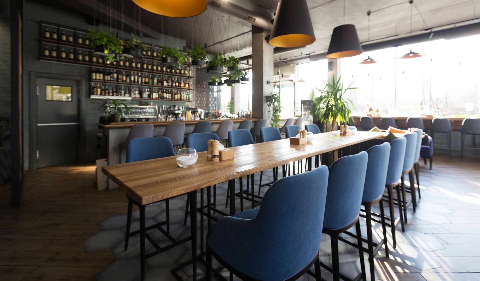 Restaurant interior with a long wooden table and blue chairs. Bar in the background with a shelf of drinks.