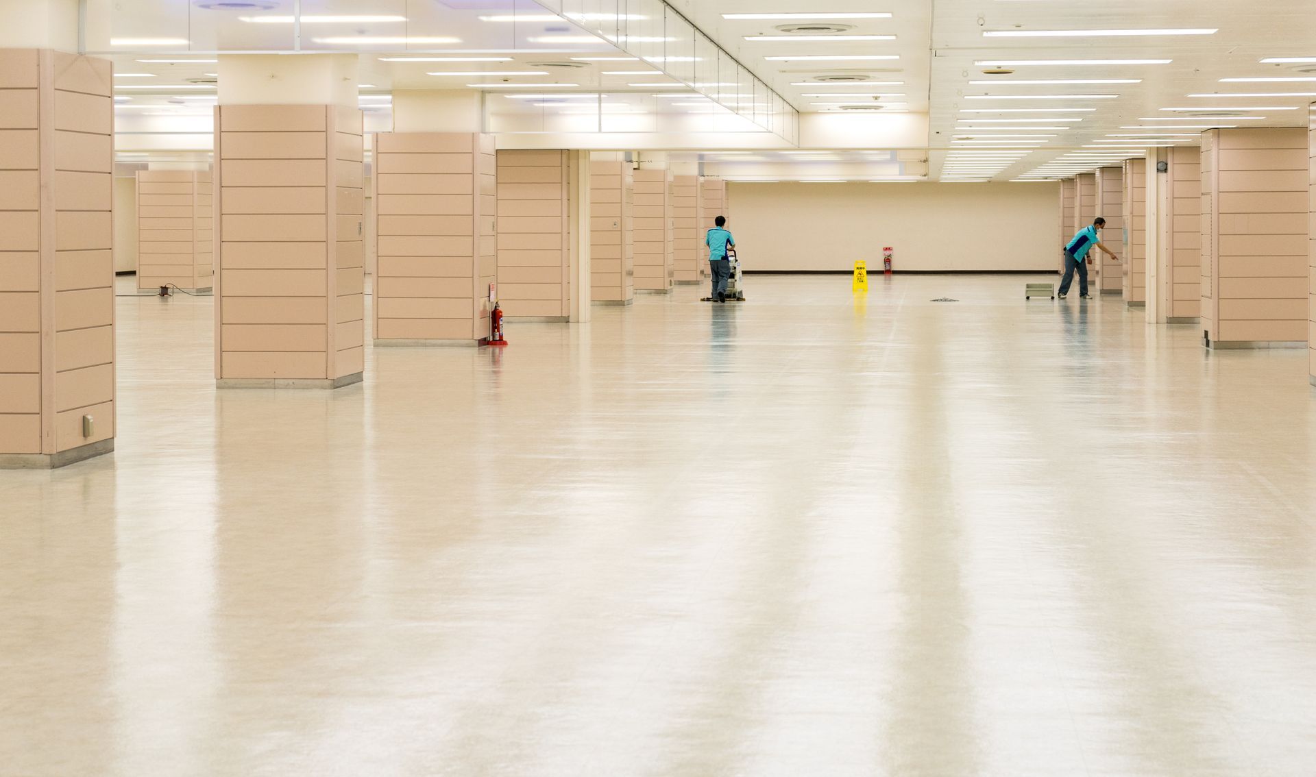 Two people cleaning a large, empty, light-colored, interior space with many support columns.