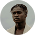 Man with dark skin and braided hair wearing a tan blazer, looking to the left.