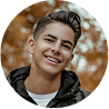 Young man with braces smiles broadly outdoors, with brown foliage in the background.