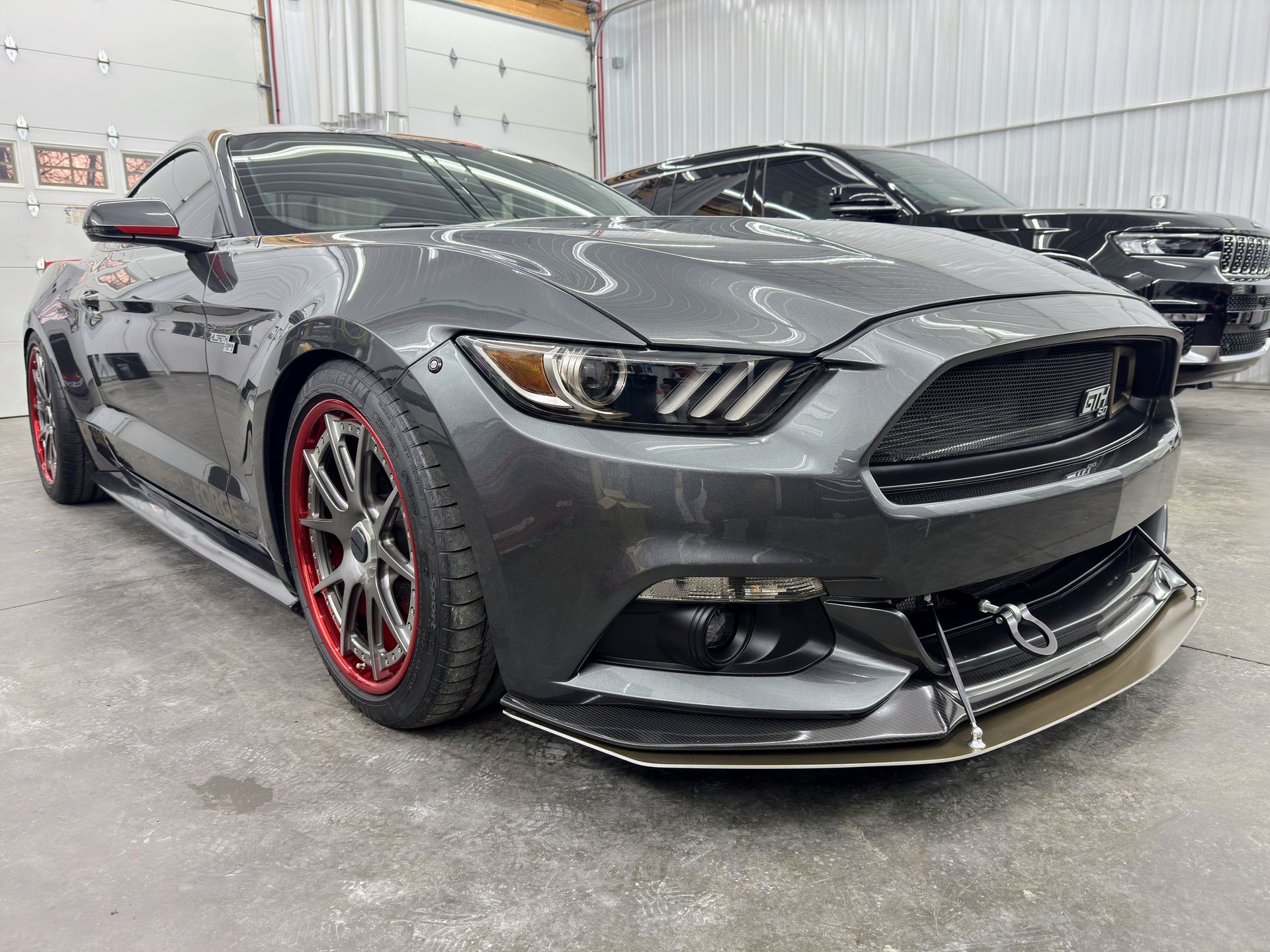 Gray sports car with red-rimmed wheels in a garage, front splitter and tow hook visible