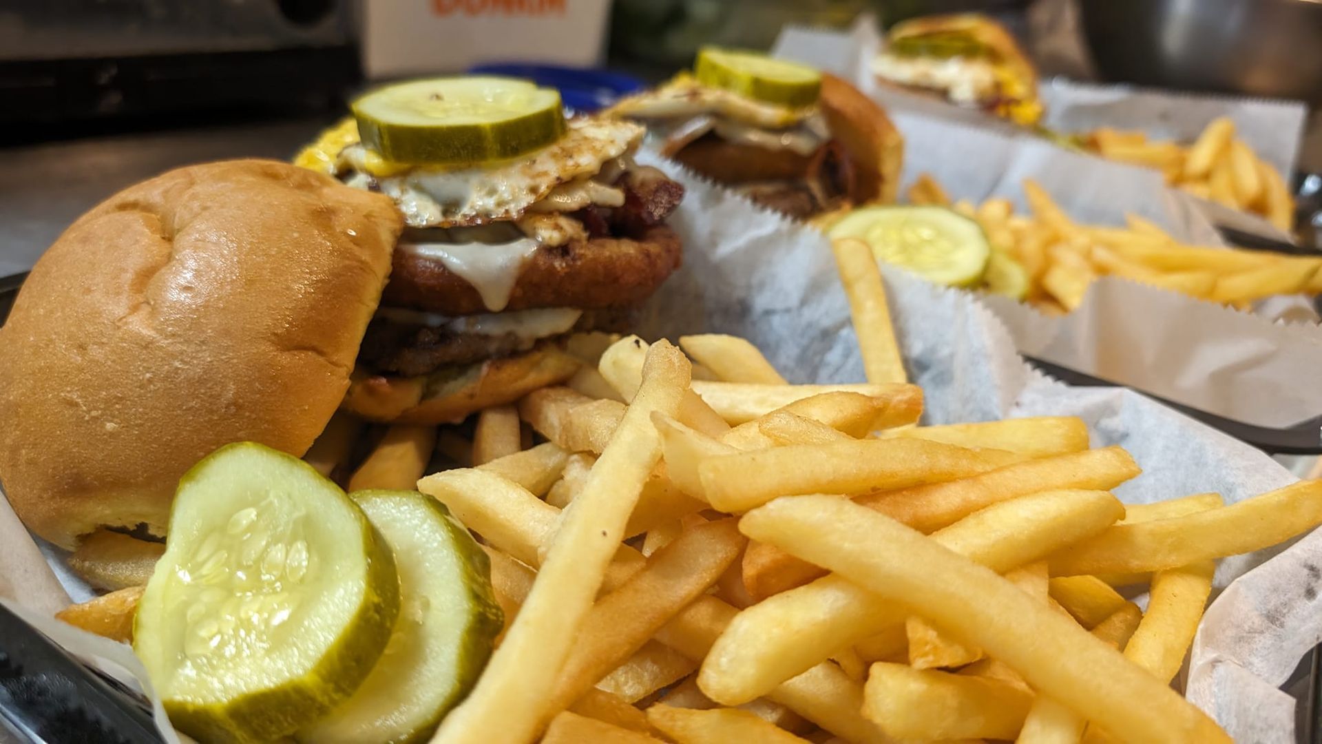 Burger and fries on a table, with pickles and burgers in the background.