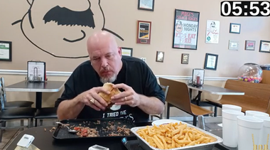 Man eating a sandwich at a diner table with fries and drinks nearby, wall decor in the background.