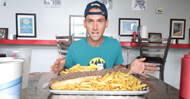 Man seated at a table with a huge tray of fries and sandwiches in a diner, smiling at the camera