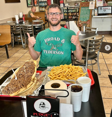 Man smiling at a restaurant table with a huge sandwich, fries, and drinks, giving two thumbs up