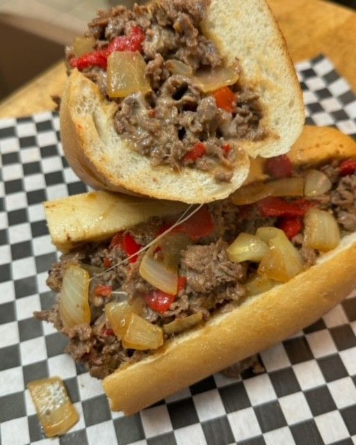 Two open cheesesteak sandwiches on checkered paper, with beef, onions, and red peppers.