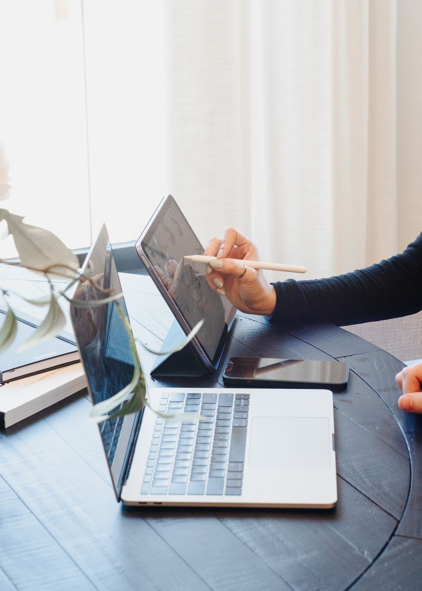 Person sketching at a laptop on a dark table beside a vase by a bright window