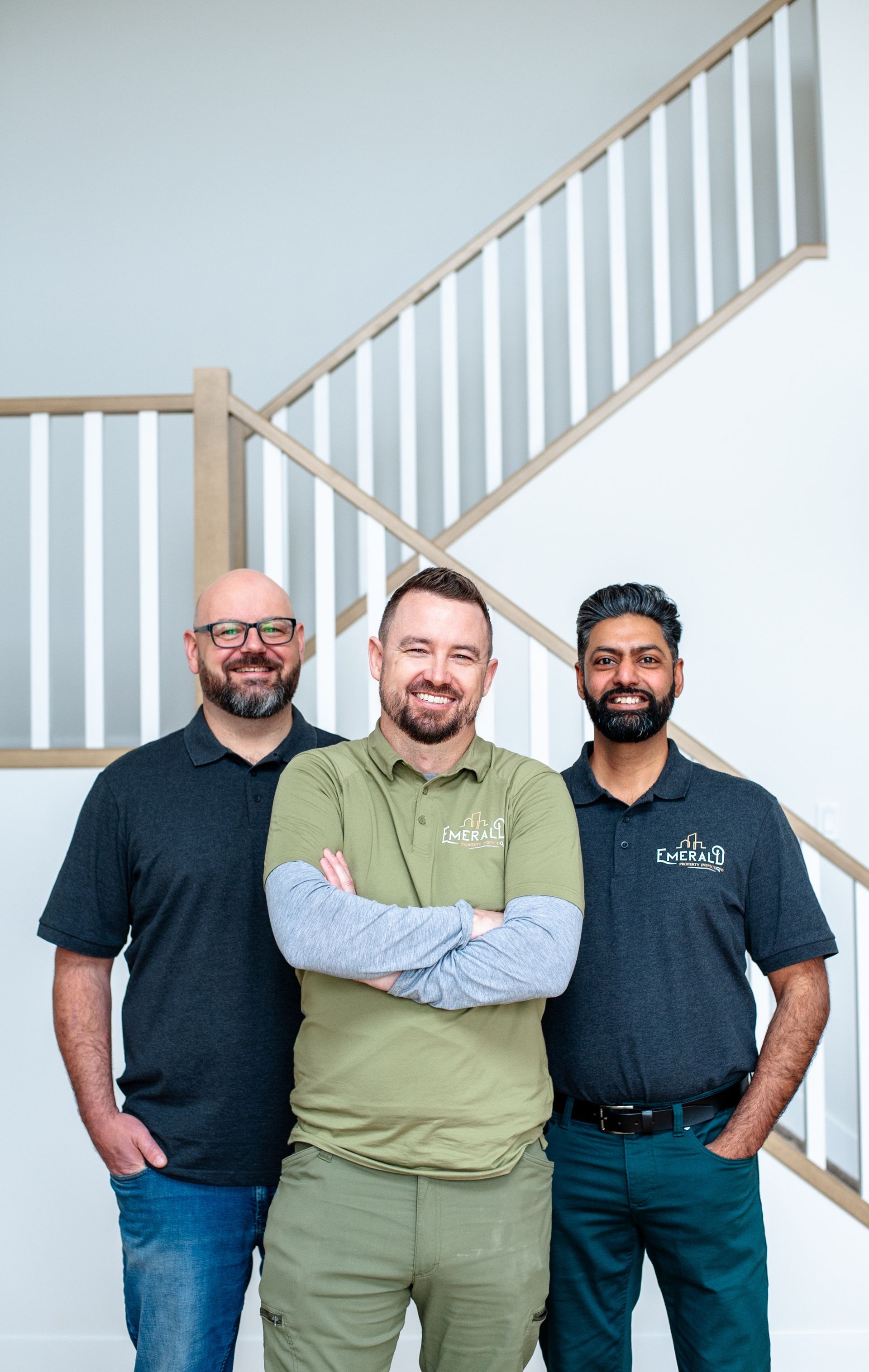 Three coworkers standing indoors by a staircase, smiling with arms crossed.