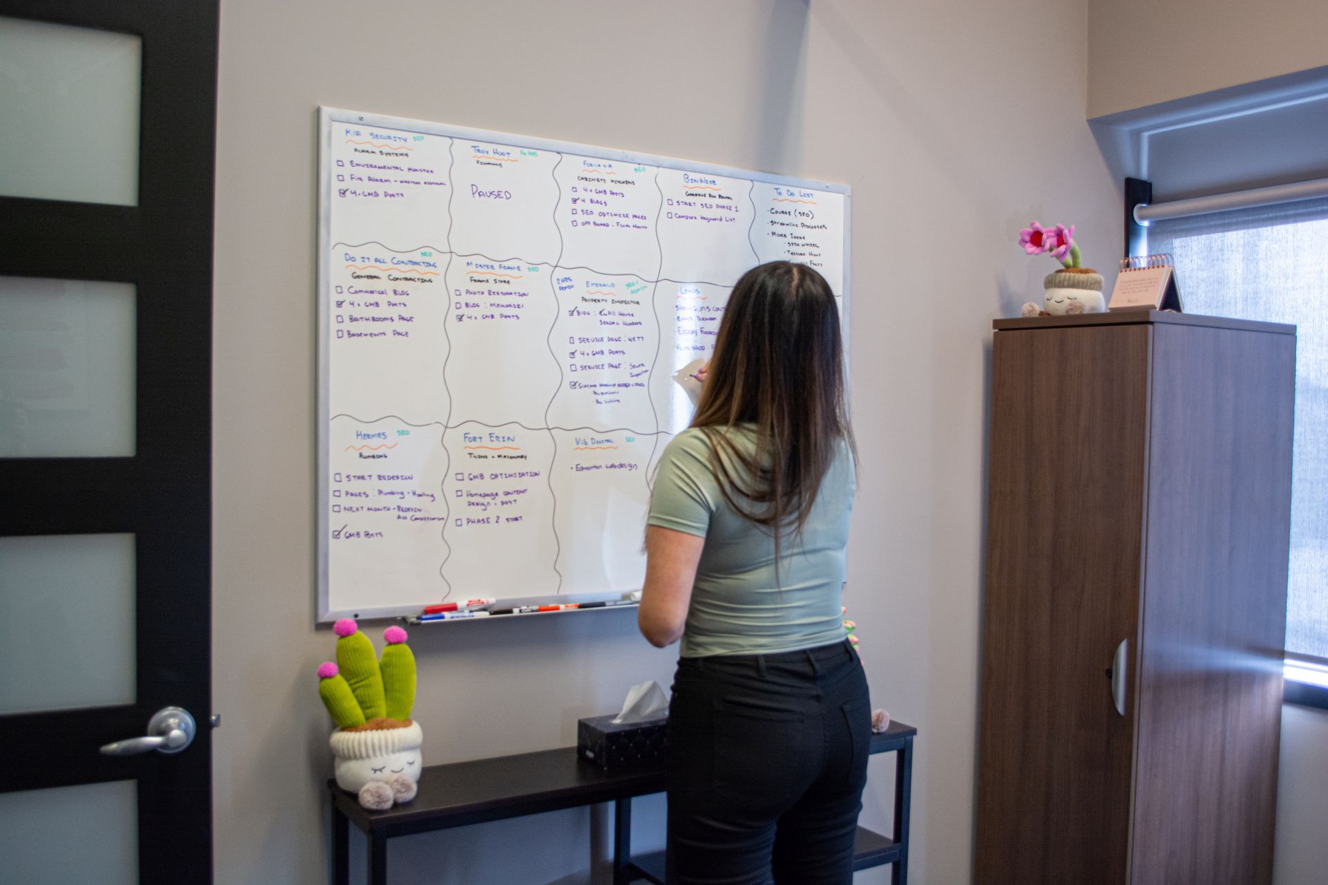 Person standing at a whiteboard in an office, writing notes near a desk and cabinet.