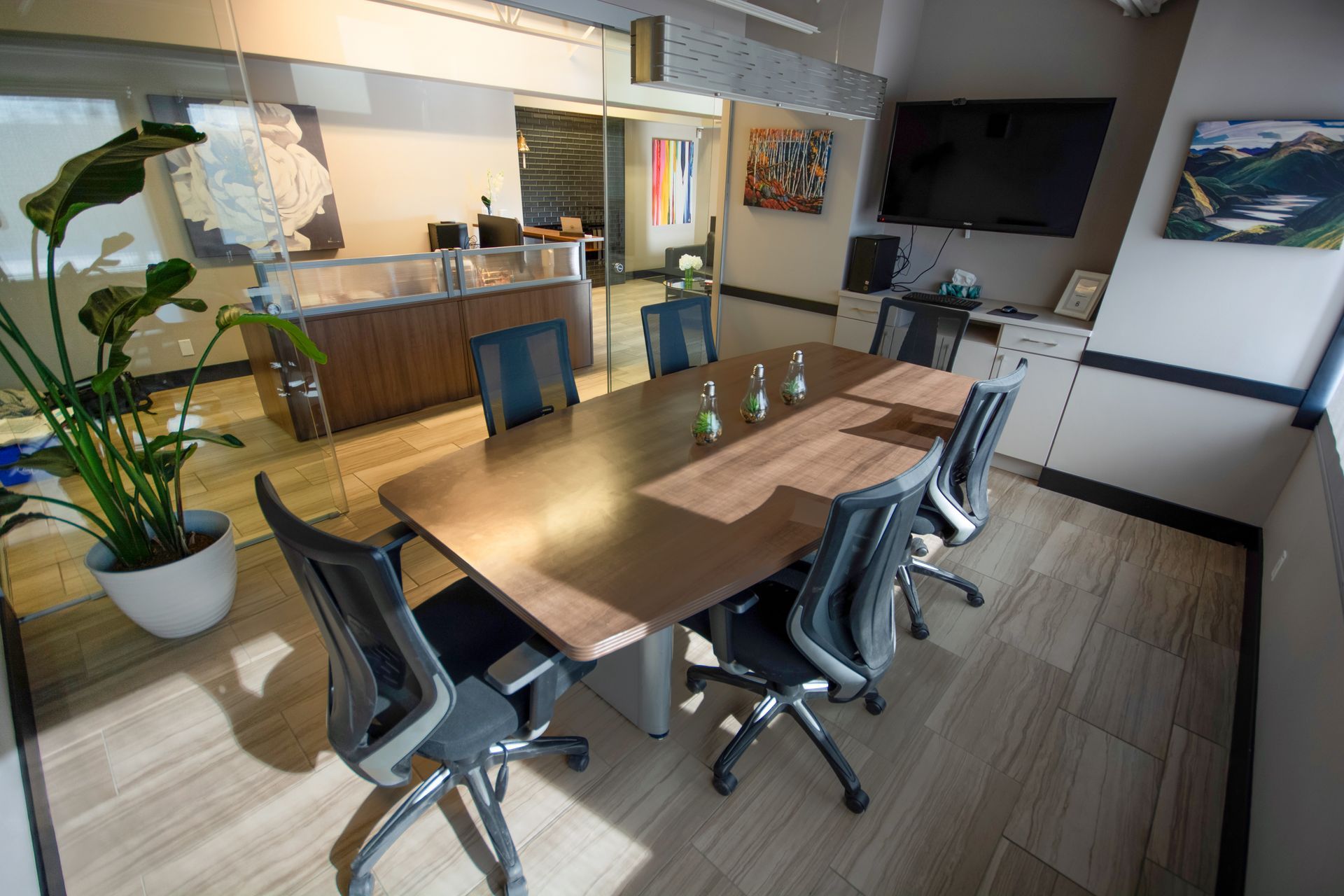 Modern conference room with long wooden table, black chairs, potted plant, and kitchen area in back