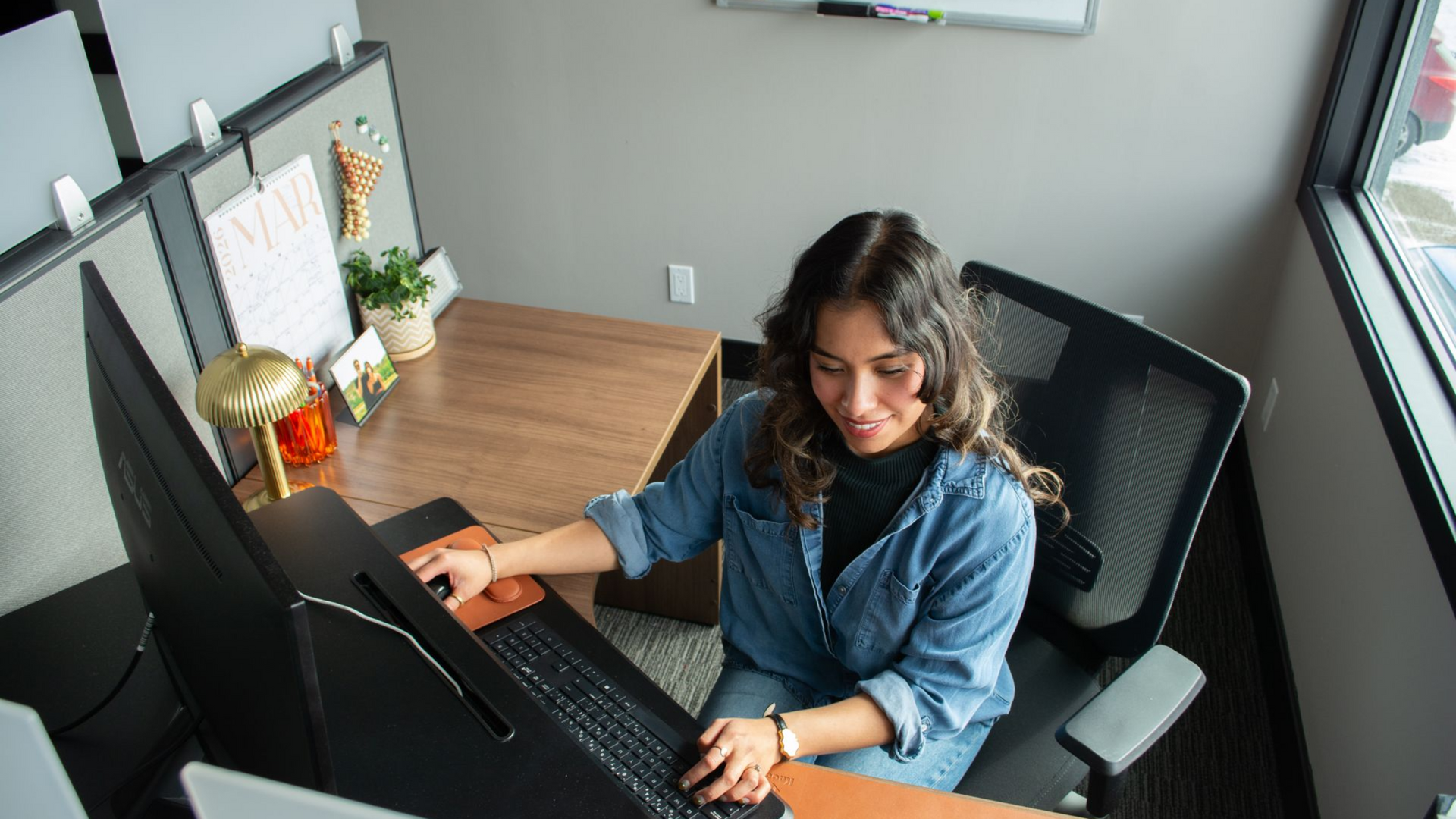 Woman working at a desk in a modern office, using a laptop and looking at a monitor.