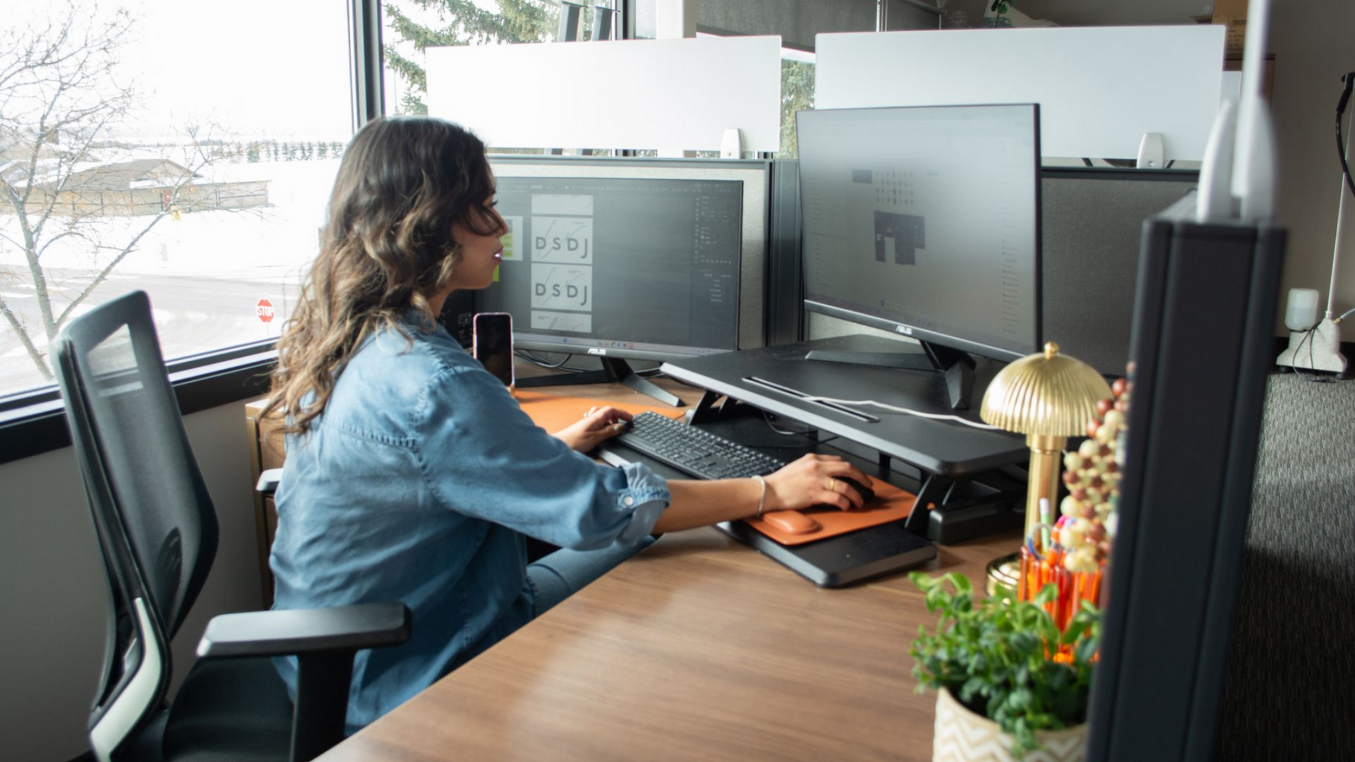 Person typing at a dual-monitor desk in a bright office, with a laptop and keyboard on a wooden workstation.