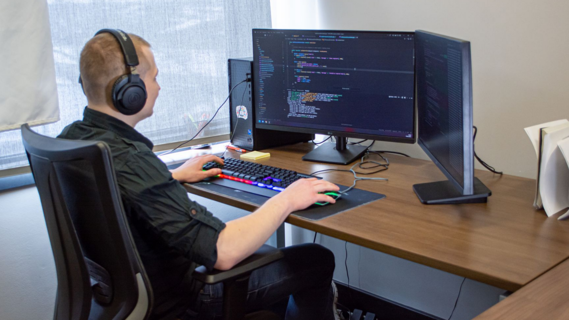 Person wearing headphones works at a desktop computer displaying code on a wooden desk.
