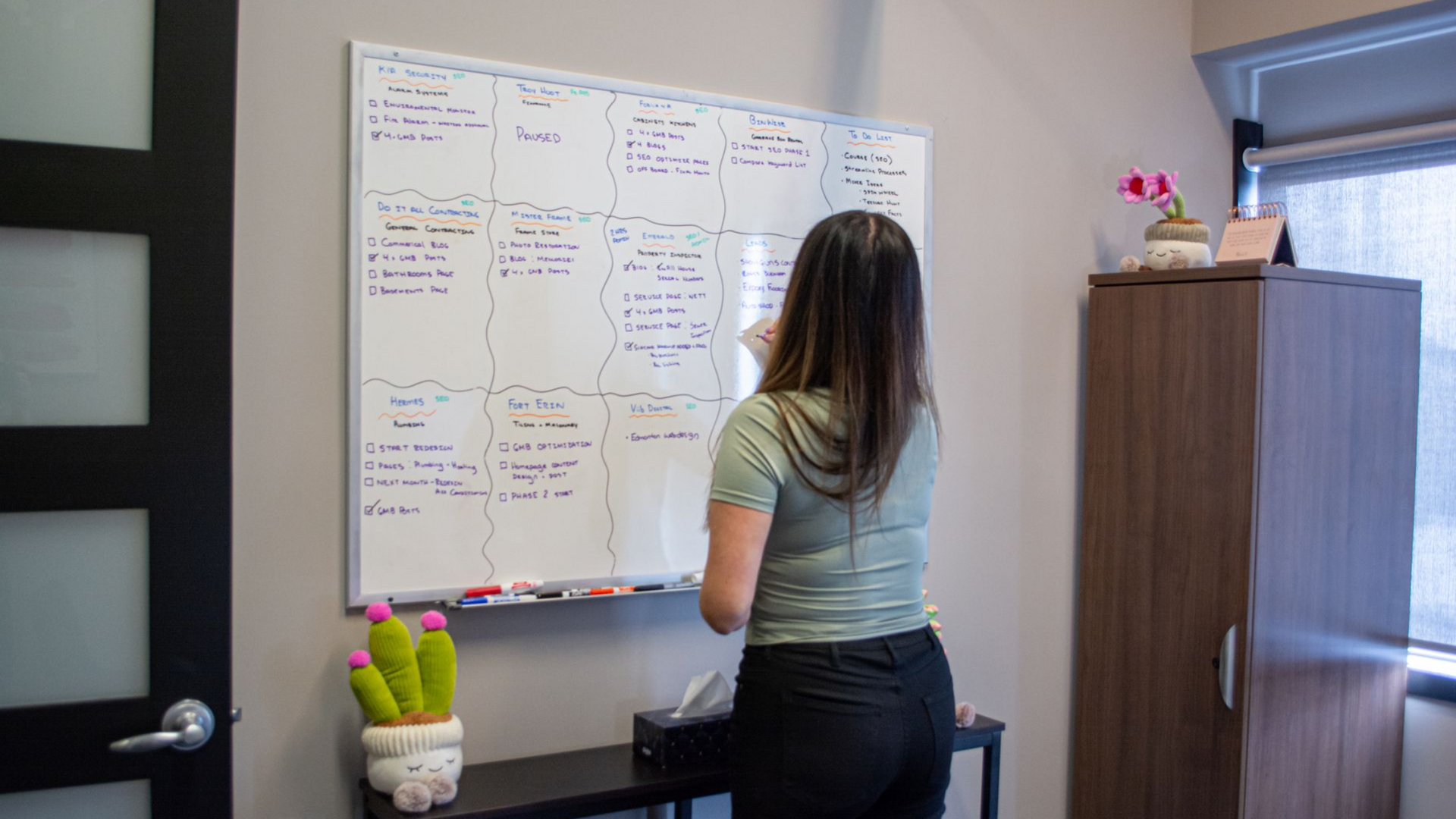 Person writing on a whiteboard in an office, with notes and a cabinet nearby.