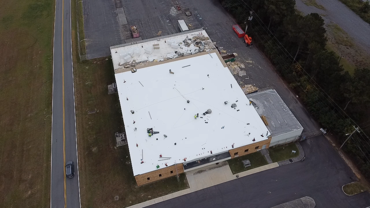 Aerial view of a rectangular building with a large white roof, situated next to a road and a parking lot with a vehicle.