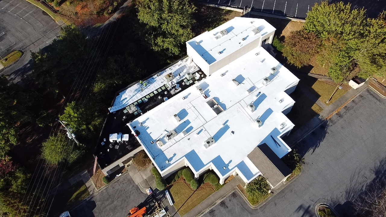 An aerial view of a building with a bright white flat roof surrounded by parking lots, trees, and landscaping.