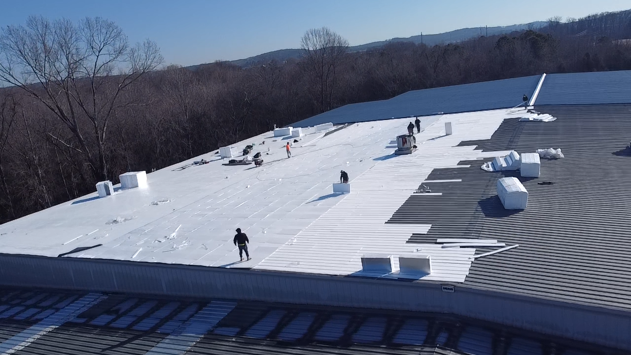 Workers install white waterproof membrane rolls on a large commercial flat roof under a clear blue sky.