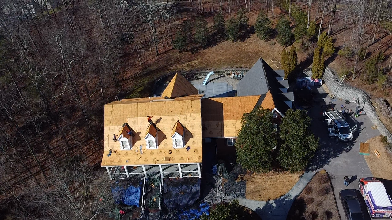 An aerial view shows a house undergoing roof renovations, with sections of exposed plywood and three dormer windows.