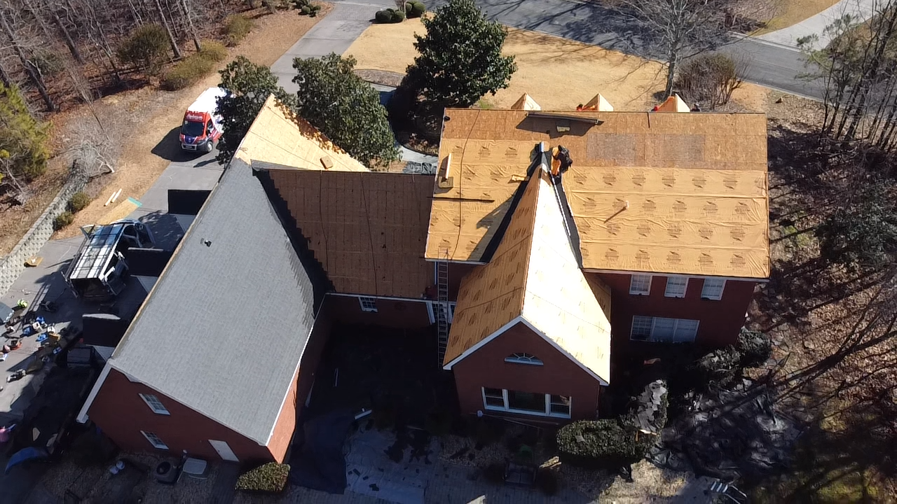 Aerial view of a house during a roof replacement, showing sections with plywood decking and others with new shingles.