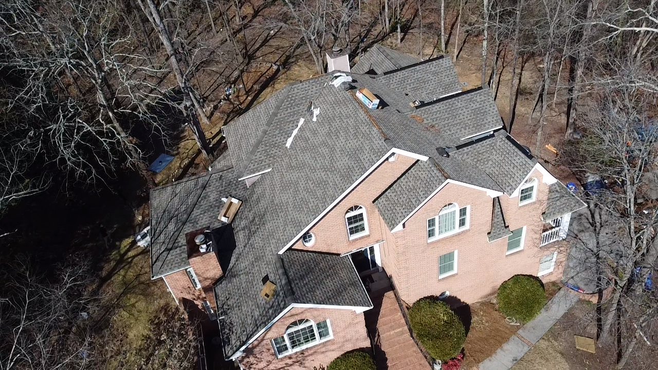 An aerial view of a multi-story brick house with a complex shingled roof, surrounded by bare trees in a wooded setting.