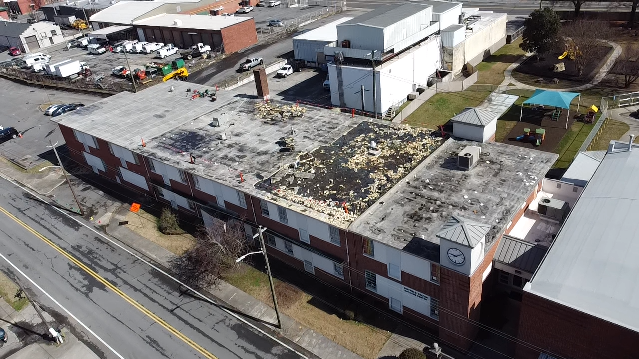 An aerial view of a rectangular, multi-story brick building with a damaged, partially covered roof, set beside a road.