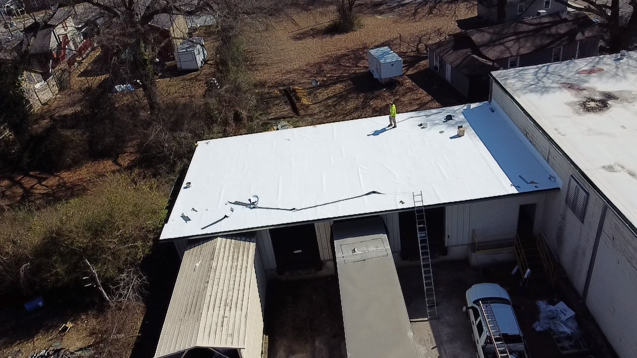 An aerial view of a person in a safety vest working on a white flat roof of a building with loading docks below.