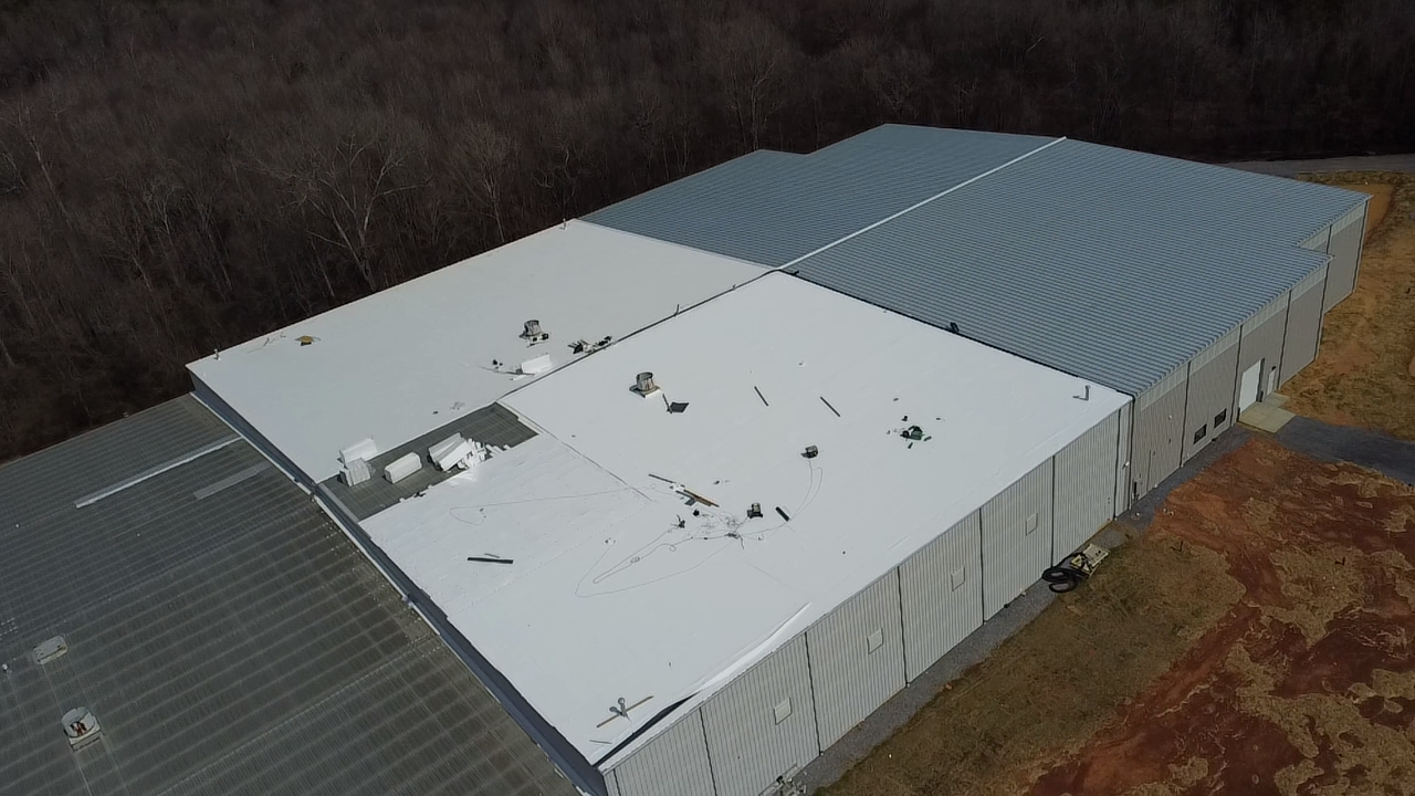 Aerial view of a large, rectangular industrial warehouse building with a light gray metal roof, surrounded by trees.