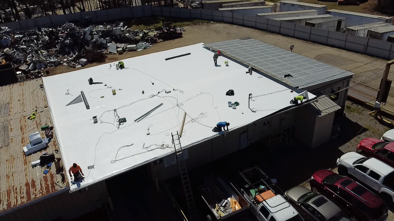 Workers install a white roof membrane on a flat building, with vehicles parked below and scrap metal in the background.