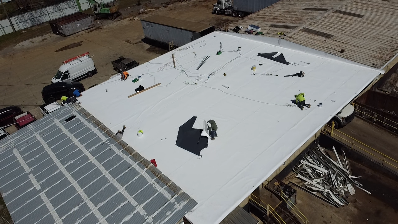 Aerial view of workers installing a white membrane roof on a building, with construction materials and vehicles nearby.