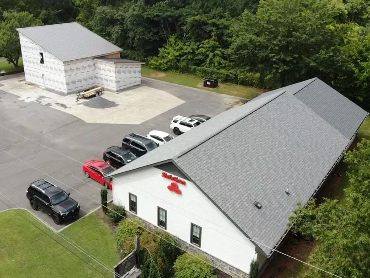 Aerial view of a white commercial building with a gray roof and parking lot, next to a building under construction.