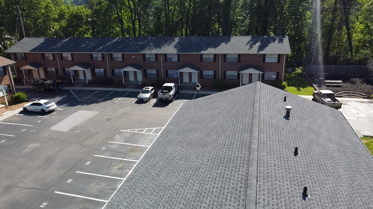 Aerial view of an apartment complex with a parking lot and a shingled roof in the foreground.