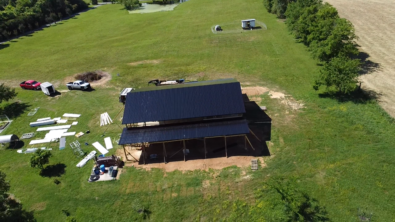 An aerial view of a dark roofed building under construction in a large grassy field with vehicles and building materials.