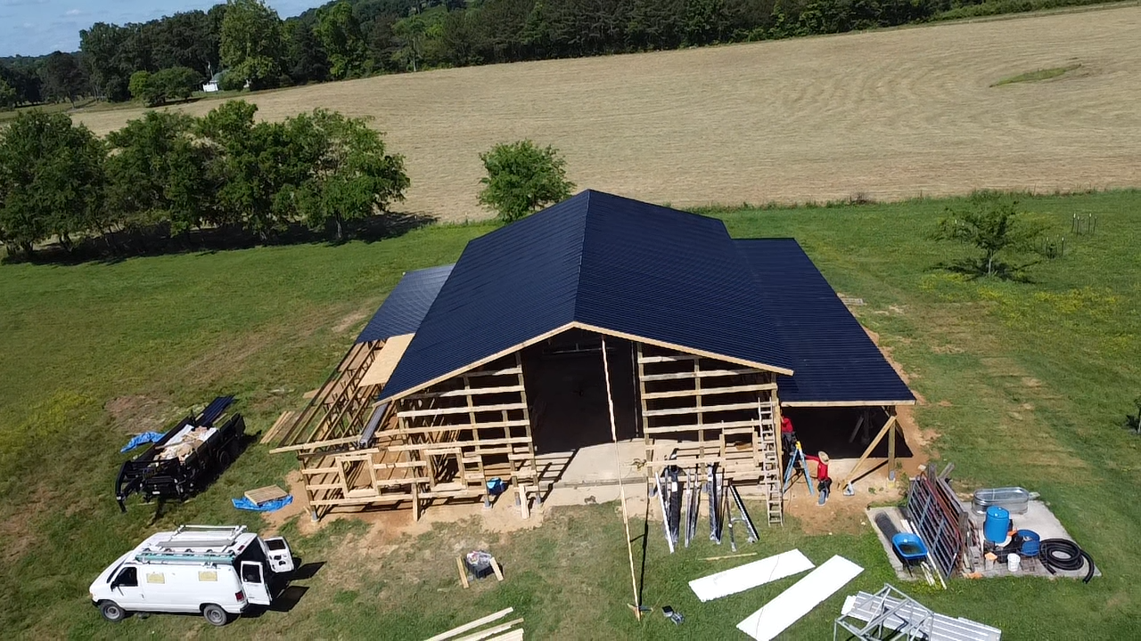 An aerial view of a wooden barn under construction in a grassy field, featuring a dark roof and workers on site.