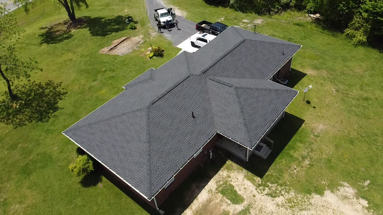 An aerial, high-angle view of a dark shingled roof on a brick house, with two vehicles parked in the adjacent driveway.