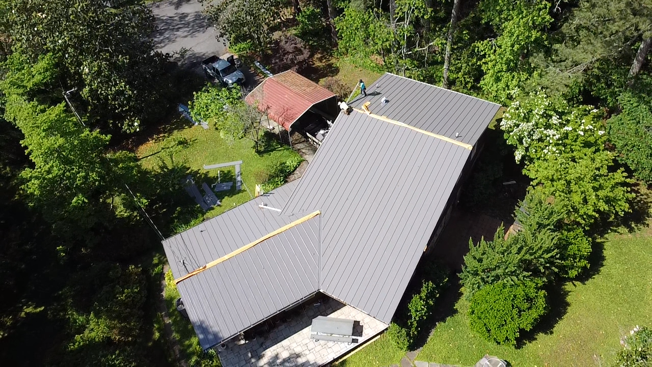 An aerial view of a gray-roofed house surrounded by trees, with a smaller reddish outbuilding nearby.