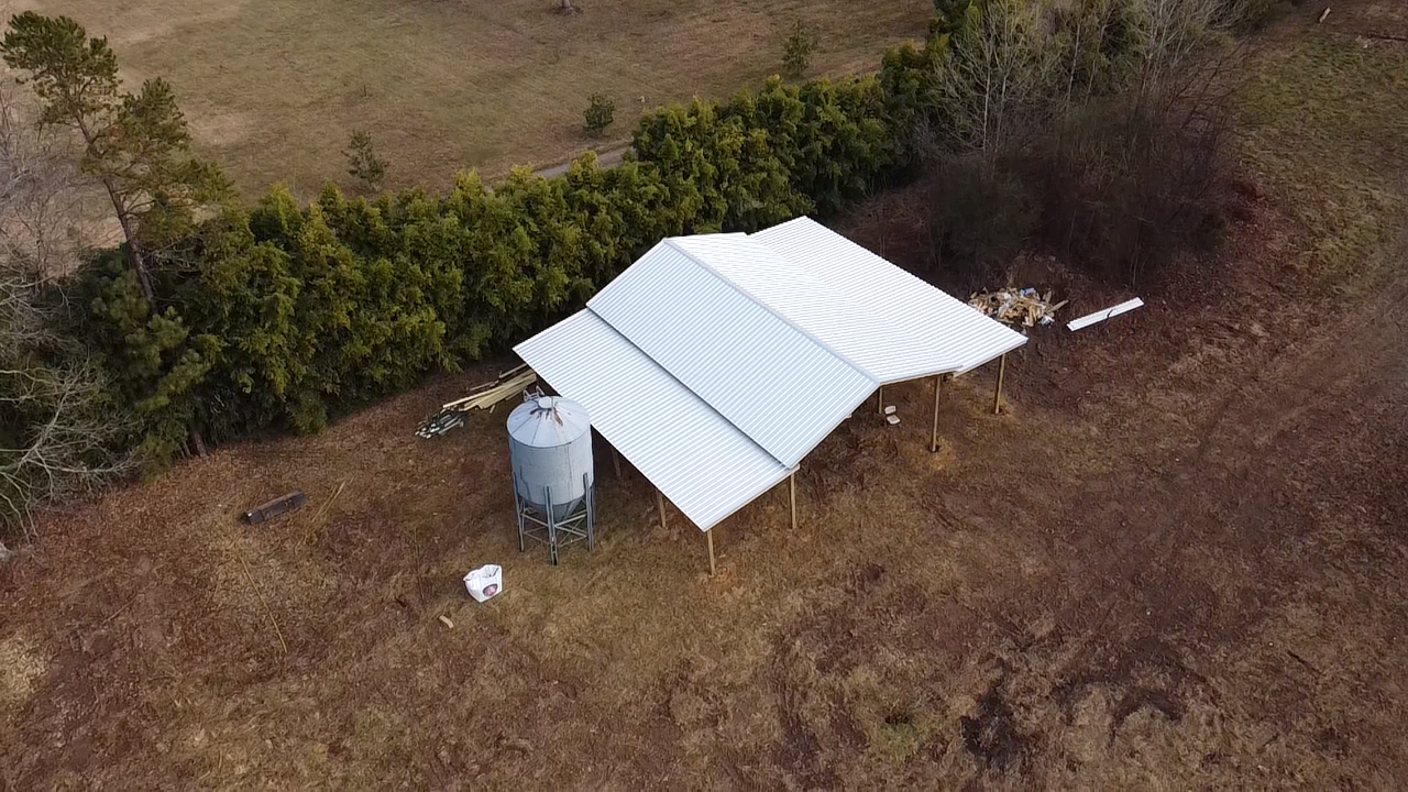 An aerial view of a metal-roofed shelter in a grassy, rural field next to a cylindrical storage bin and a line of trees.