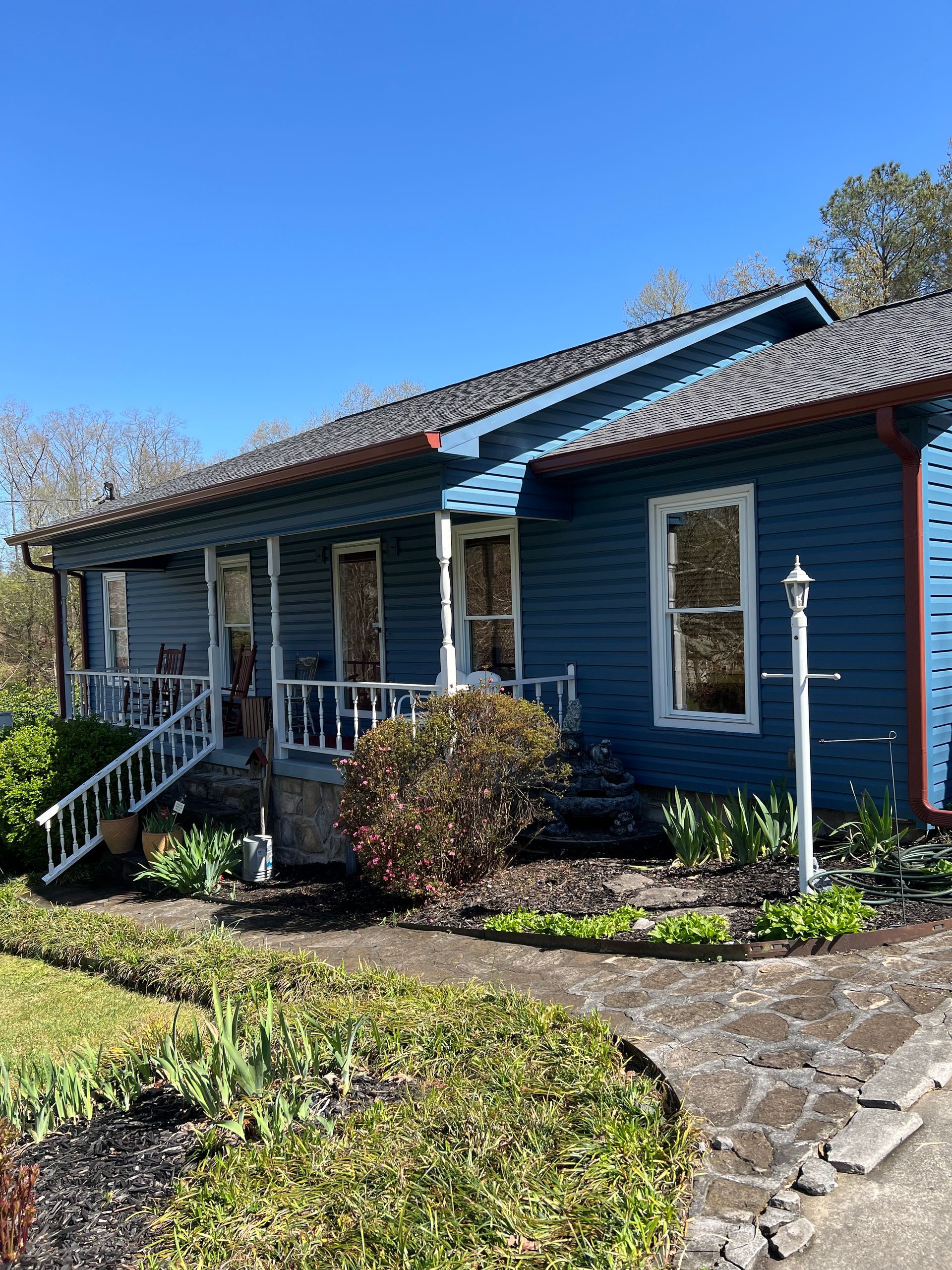 A blue wooden house with a front porch, white railings, and a landscaped yard under a clear blue sky.