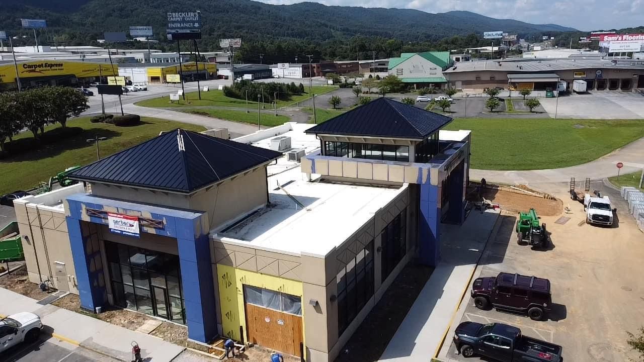 An aerial view of a commercial building under construction with blue accents, dark metal roofs, and surrounding parking.