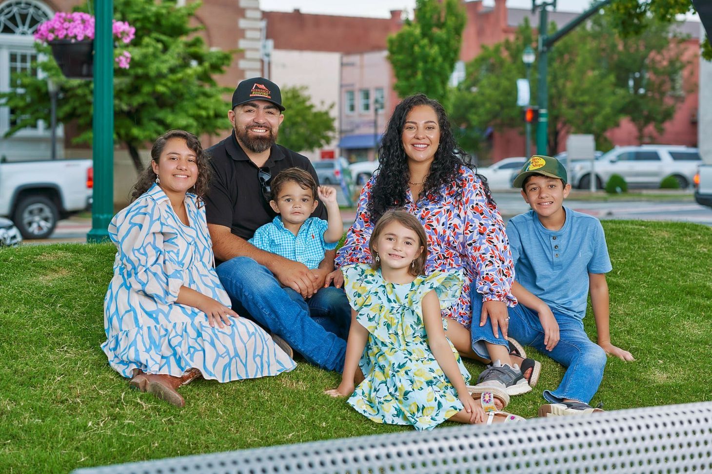 A family of six poses for a portrait on a grassy patch in a town square, smiling toward the camera.
