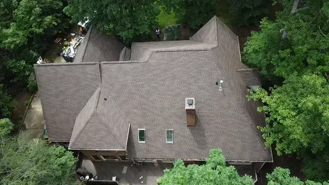 An aerial view of a multi-gabled brown shingle roof on a house surrounded by trees.