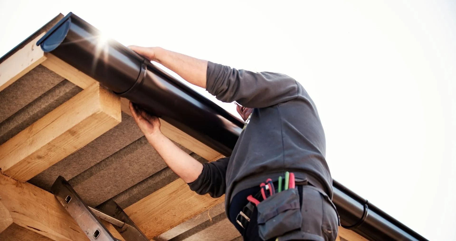 A worker wearing a tool belt installs a dark-colored gutter onto the wooden eaves of a roof.